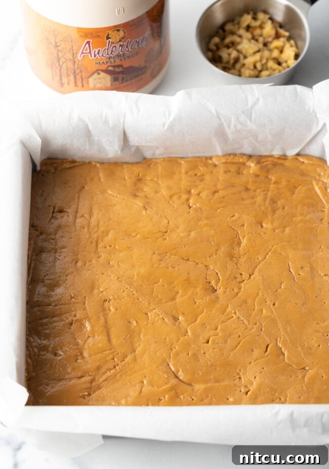 Smooth maple fudge mixture pressed firmly into a square baking pan lined with parchment paper, ready for chilling.