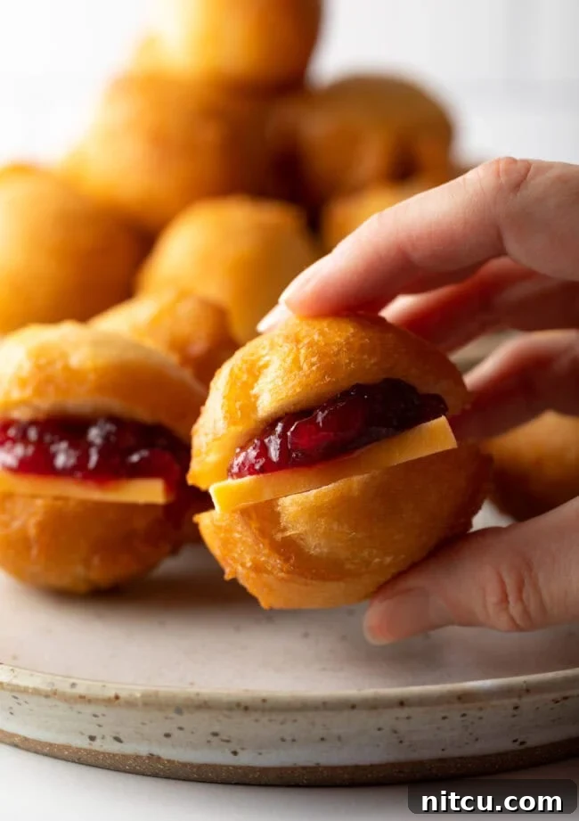A hand holding a small Vetkoek (fat cake) fried dough, cut open and filled with vibrant cheese and jam.