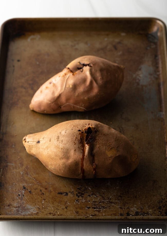 Two perfectly baked sweet potatoes, still in their skins, resting on a baking sheet, ready to be cooled and mashed.