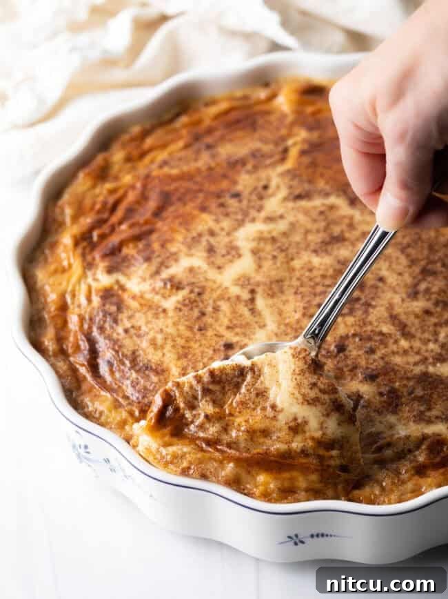 A hand scooping a generous serving of baked rice pudding from a large rectangular baking dish using a metal spoon.