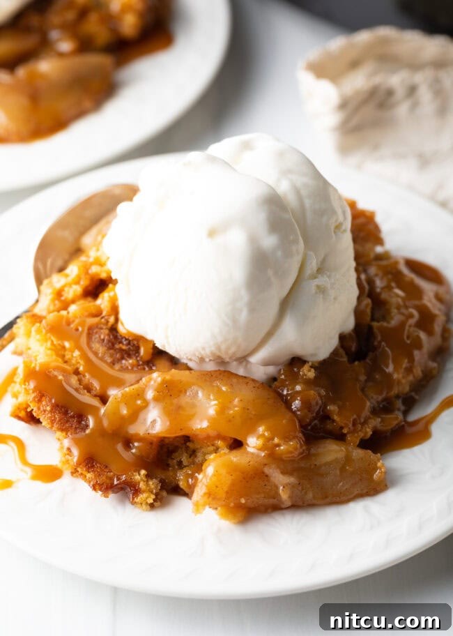 A close-up view of a slice of warm Caramel Apple Dump Cake on a white plate, elegantly served with a scoop of creamy vanilla ice cream.