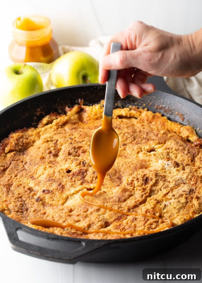 A hand holds a spoon, artfully adding a stream of thick, luscious caramel sauce over the warm, baked skillet of apple dump cake.