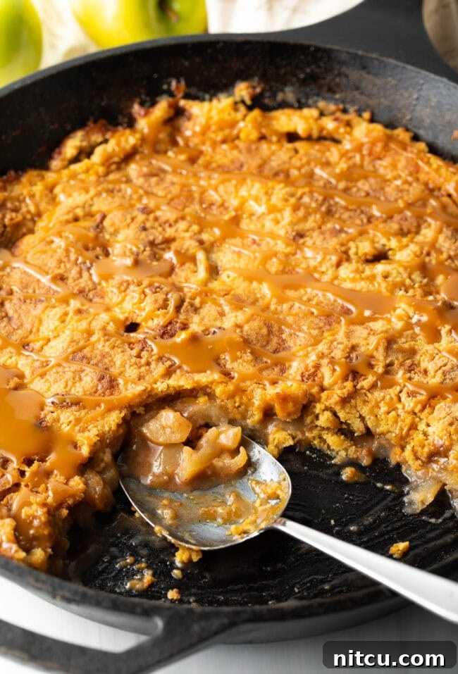 A close-up view of the baked Caramel Apple Dump Cake in a cast-iron skillet, with a serving spoon removing a portion, highlighting its soft, gooey texture.