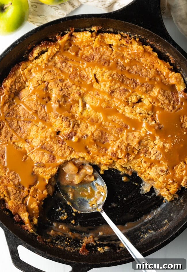 A top-down view of a golden-brown Caramel Apple Dump Cake baked perfectly in a cast-iron skillet, with a portion already removed by a metal spoon, showcasing its gooey center.