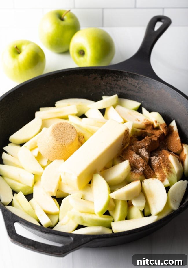 Freshly sliced Granny Smith apples, a stick of unsalted butter, rich brown sugar, and aromatic spices ready in a cast iron skillet for the apple filling.