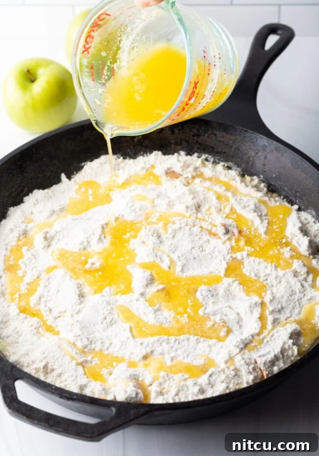 Melted butter being carefully poured from a glass measuring cup, evenly coating the dry box cake mix layer atop the apple filling.