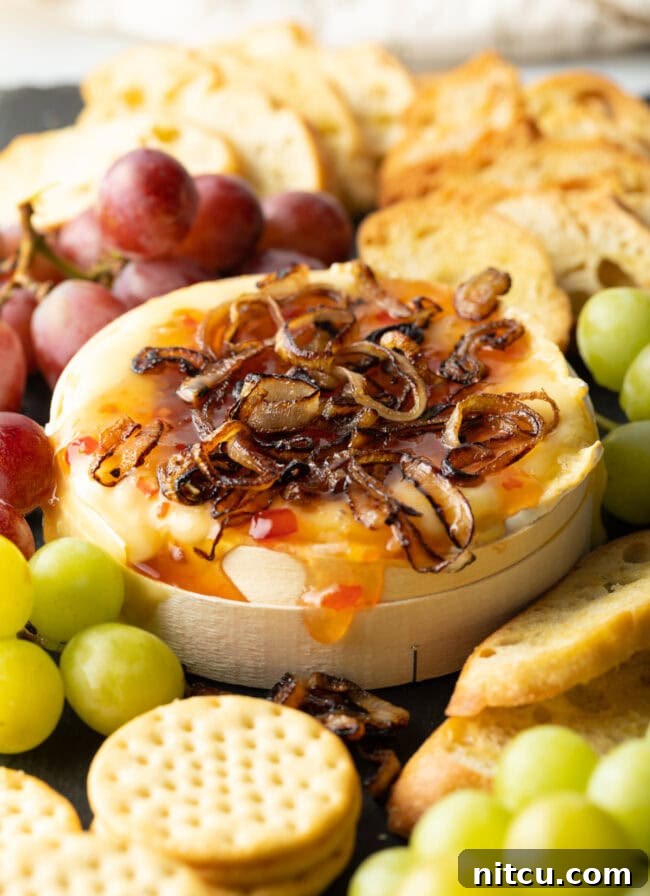 Wheel of Camembert cheese baked with sweet chili sauce and sauteed shallots. It is on a platter and surrounded by red and green grapes, crackers, and baguette slices.