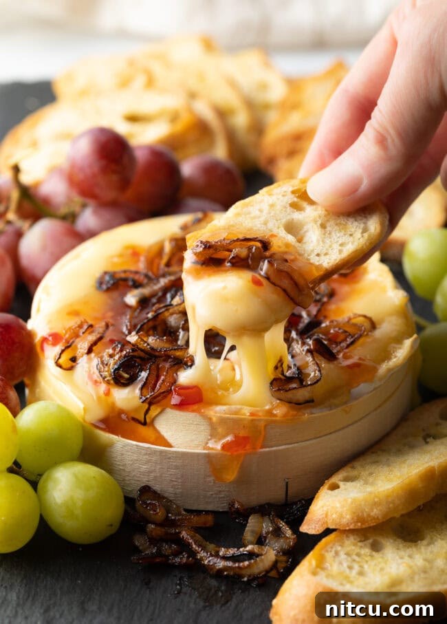 Hand scooping gooey melted cheese with onions and chili sauce from the baked wheel.