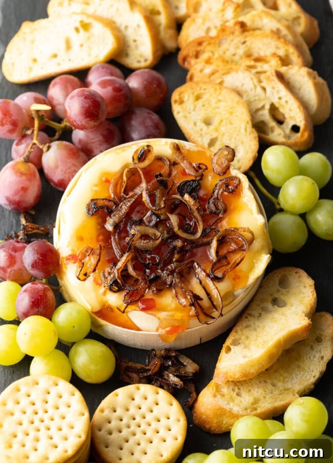 Top down view baked Camembert topped with sweet chili sauce and sauteed shallots. It is on a platter and surrounded by red and green grapes, crackers, and baguette slices.