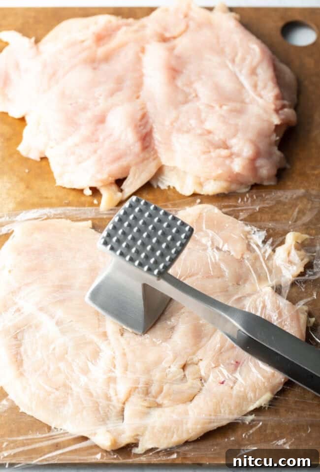 Butterflied chicken breasts on a cutting board, covered in plastic and with a metal mallet pounding them thin.