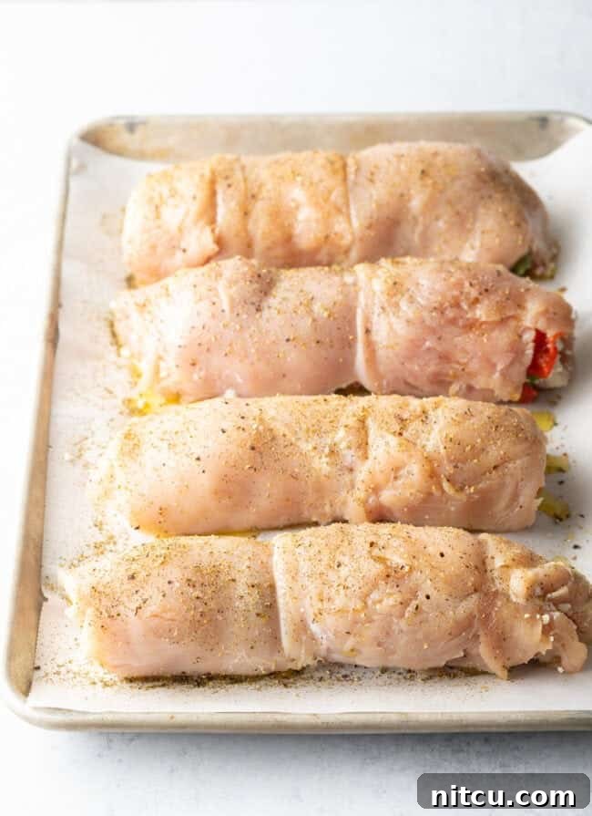 Four stuffed and rolled chicken breasts on a baking tray. They have been seasoned with olive oil and Italian seasonings, ready for the oven.