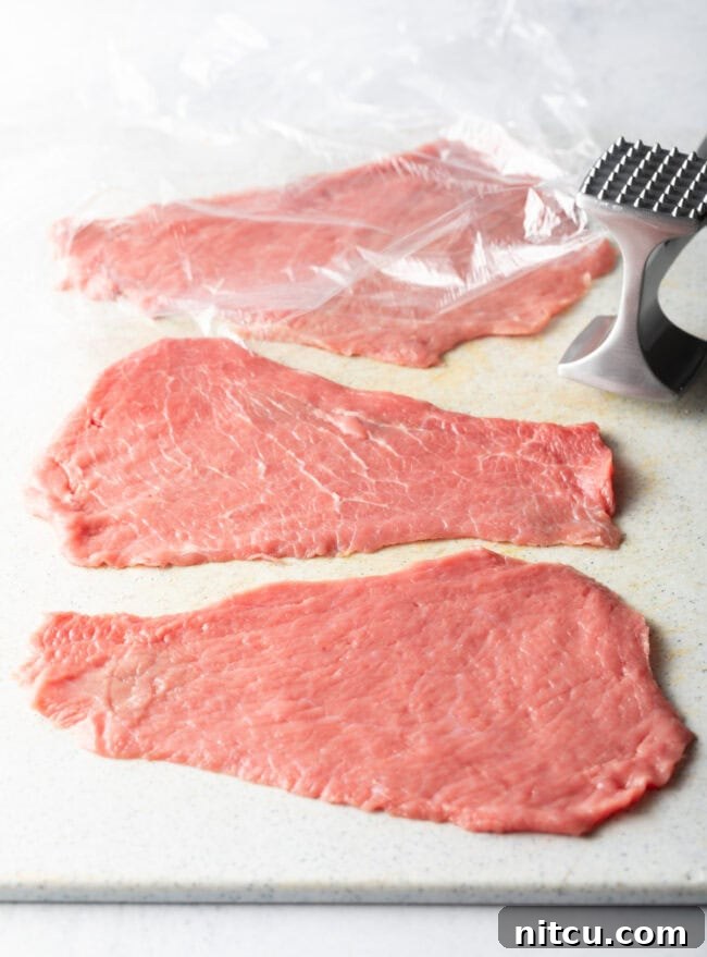 Four raw cutlets with plastic wrap and meat mallet. 