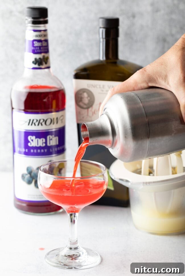 A hand pouring the vibrant pink liquid from a metal cocktail shaker into an elegant martini-style glass, illustrating the final step of preparing a Sloe Gin Fizz.