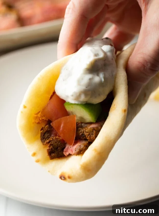 Close view hand holding flatbread naan beef shawarma recipe topped with chopped cucumbers, tomatoes, red onion and tzatziki sauce.