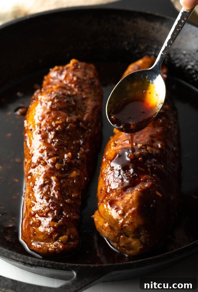 A hand spooning rich honey garlic glaze over two glistening baked pork tenderloins in a cast iron skillet.