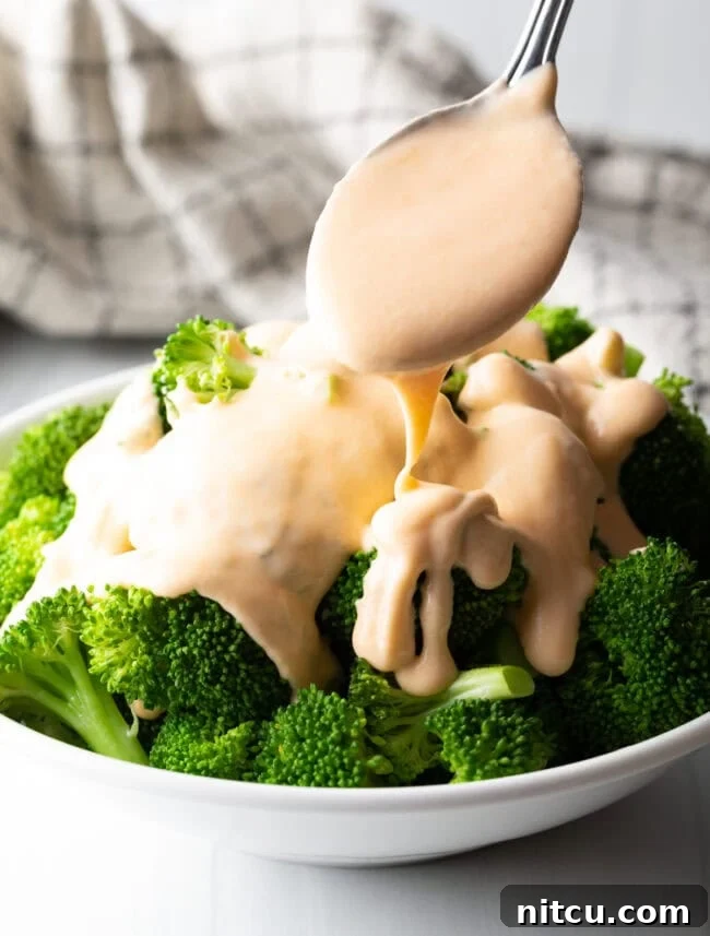 Bowl of steamed broccoli with spoon pouring cheese sauce for broccoli on top.