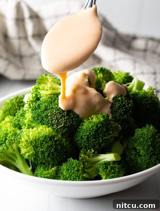Metal spoon with cheese sauce pouring over steamed broccoli.