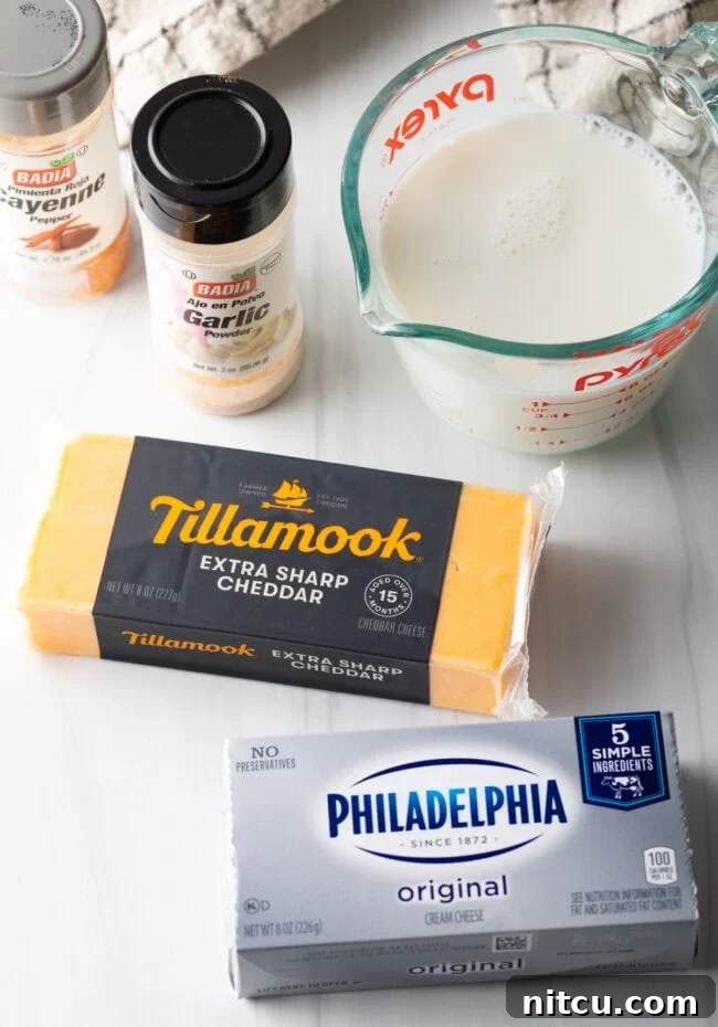 Recipe ingredients on a white board: block of sharp cheddar cheese, block of cream cheese, glass measuring pot with milk, garlic and cayenne spice bottles.