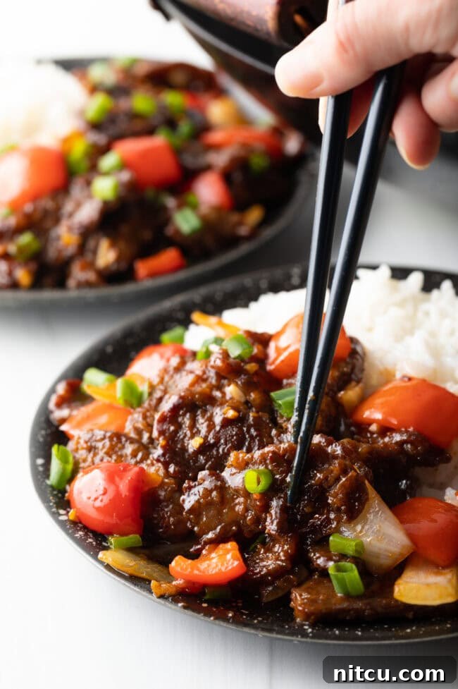 Hand with black chopsticks selecting piece of beef from a bowl of stir fry with rice.