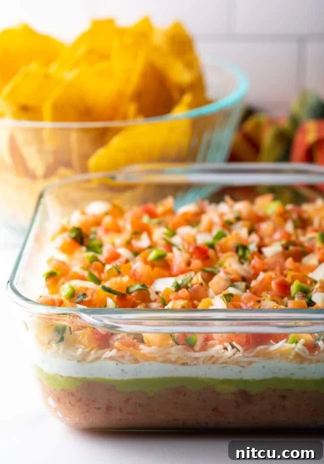 Glass square baking dish with layered Mexican bean dip. A bowl of tortilla chips is in the background.