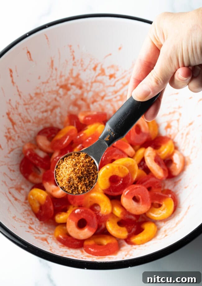 Hand adding Tajin seasoning from a metal measuring spoon into a bowl of gummy rings coated in chamoy.