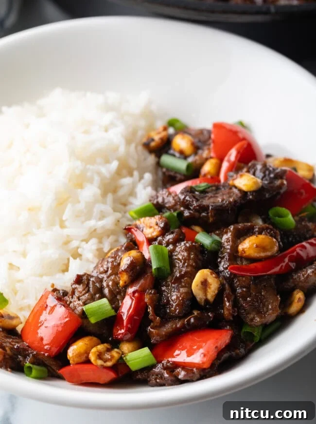 Kung pao beef stir fry serving in a white bowl with white rice, garnished with peanuts and green onions.