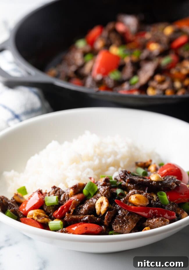 Kung pao beef stir fry serving in a white bowl with white rice, garnished with green onions.
