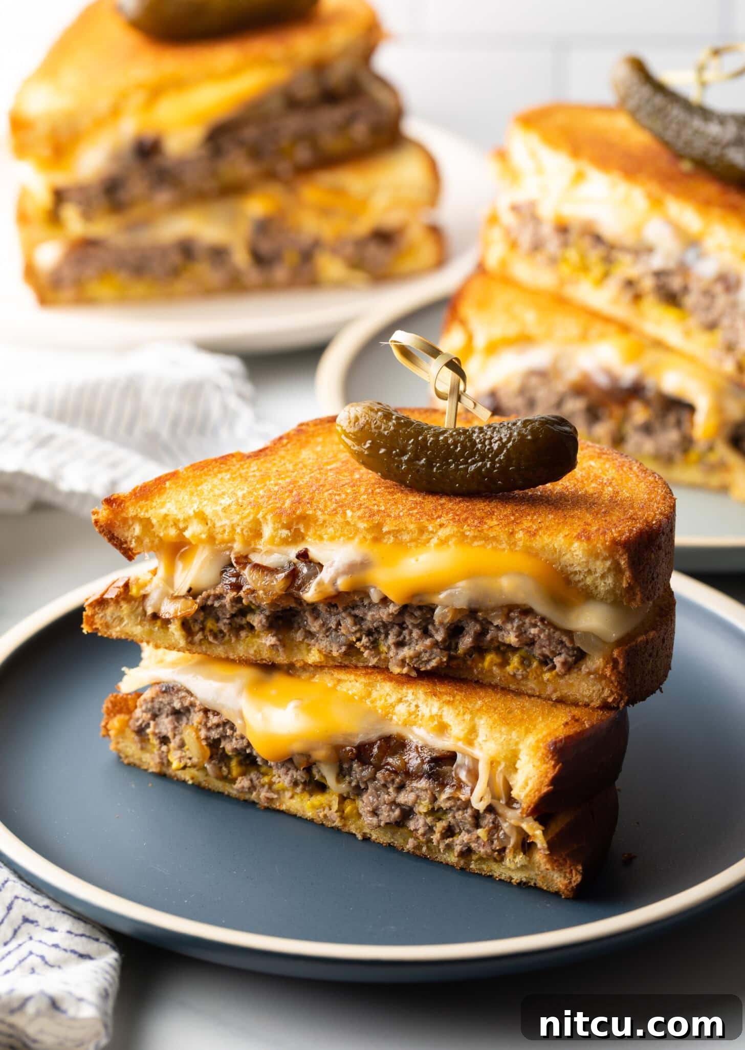 Two halves of a patty melt stacked on top of each other. A wood skewer with small pickle is going through the top of the sandwich stack.