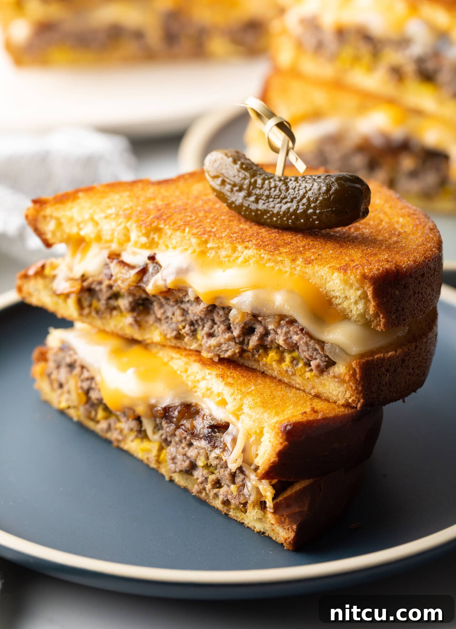Close view of two halves of a patty melt stacked on top of each other. A wood skewer with small pickle is going through the top of the sandwich stack.