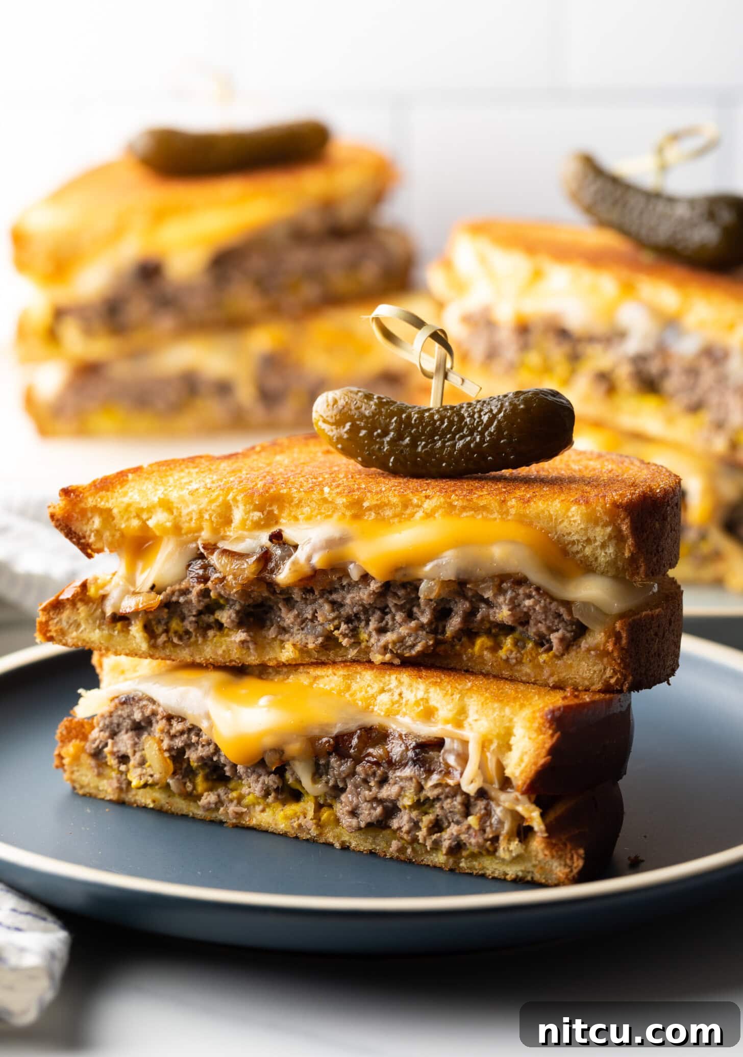 Two halves of a patty melt stacked on top of each other. A wood skewer with small pickle is going through the top of the sandwich stack.
