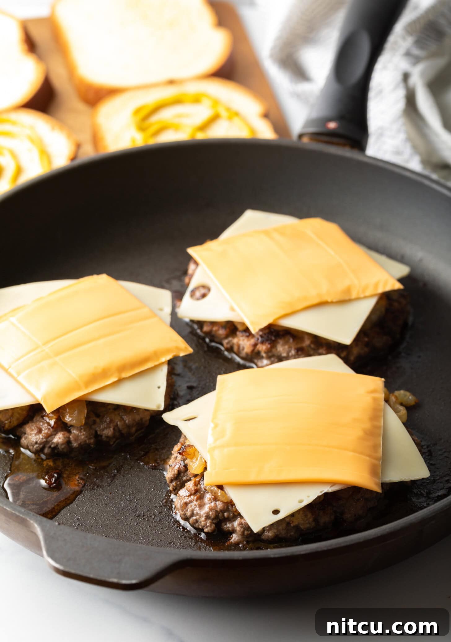 Three burger patties in a skillet with a slice of swiss cheese and slice of cheddar cheese on each.