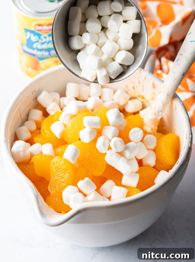 Adding mini marshmallows to the white mixing bowl with fluffy and oranges.