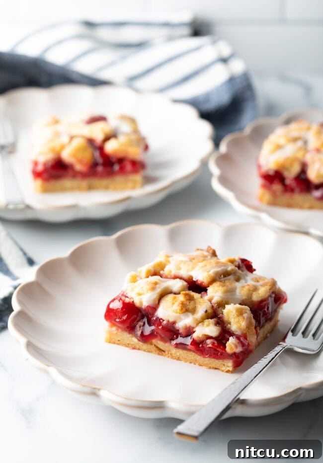 Three individual pieces of cherry pie bars, glistening with a sugar glaze, artfully arranged on white scalloped plates.