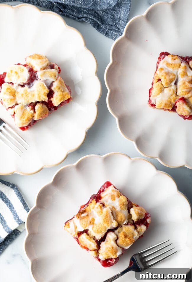 Three pieces of cherry pie bars arranged individually on elegant white scalloped plates, highlighting their golden crust and vibrant cherry filling.