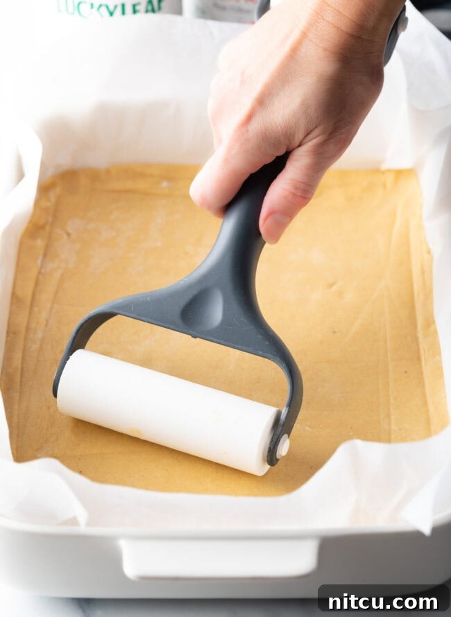 A hand uses a pastry roller to smooth out a rectangle of pie dough into an even layer in a baking pan, creating the base for the bars.