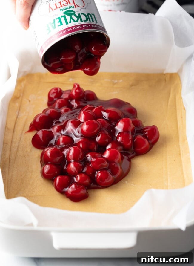 A can of vibrant red cherry pie filling is being added to a rectangular baking dish already lined with the prepared dough crust, ready for baking.