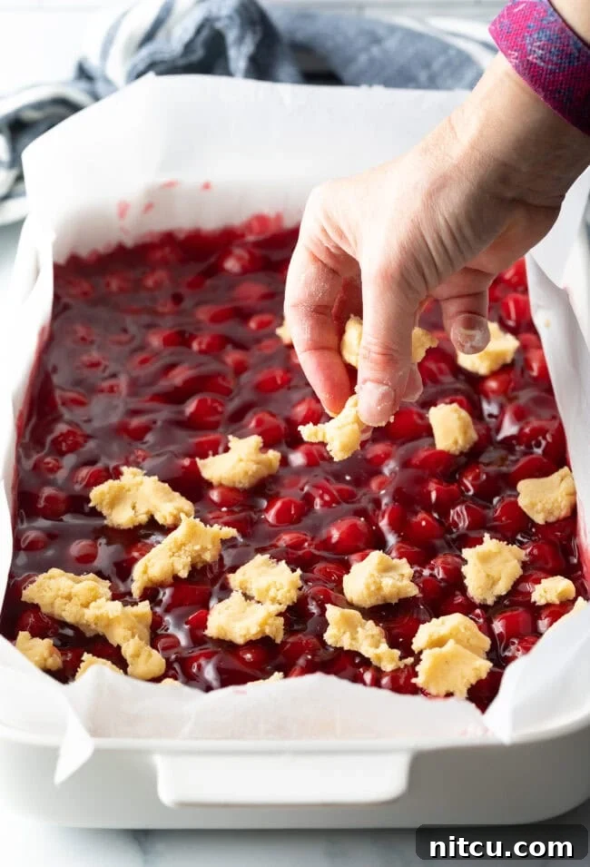 A hand carefully adds small, raw dough pieces onto the surface of the cherry pie filling, creating a rustic topping for the bars.