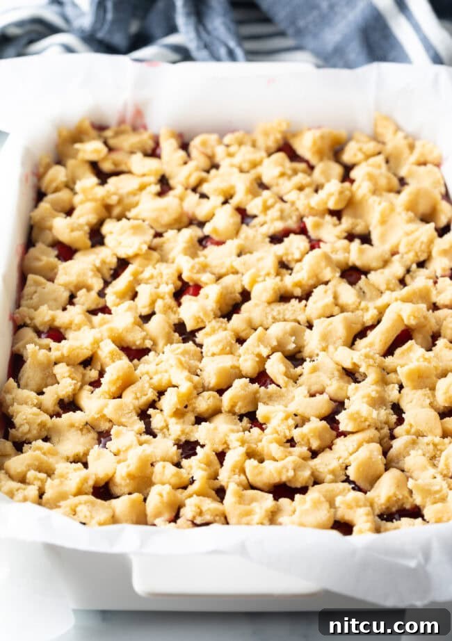 A baking pan filled with bright red cherry filling and adorned with small, raw pieces of pie dough on top, ready for the oven.