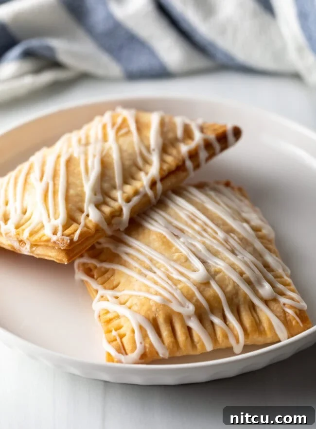 Two golden-brown homemade peach pop tarts resting against each other on a white plate, glistening with sweet icing.