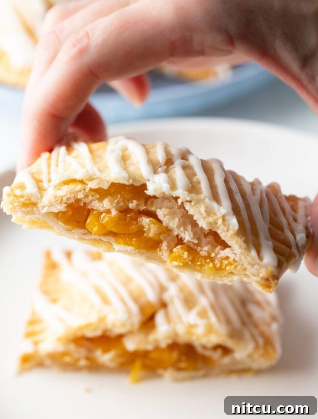 A hand holding half of a homemade peach hand pie towards the camera, showcasing the delicious, fruit-filled interior.