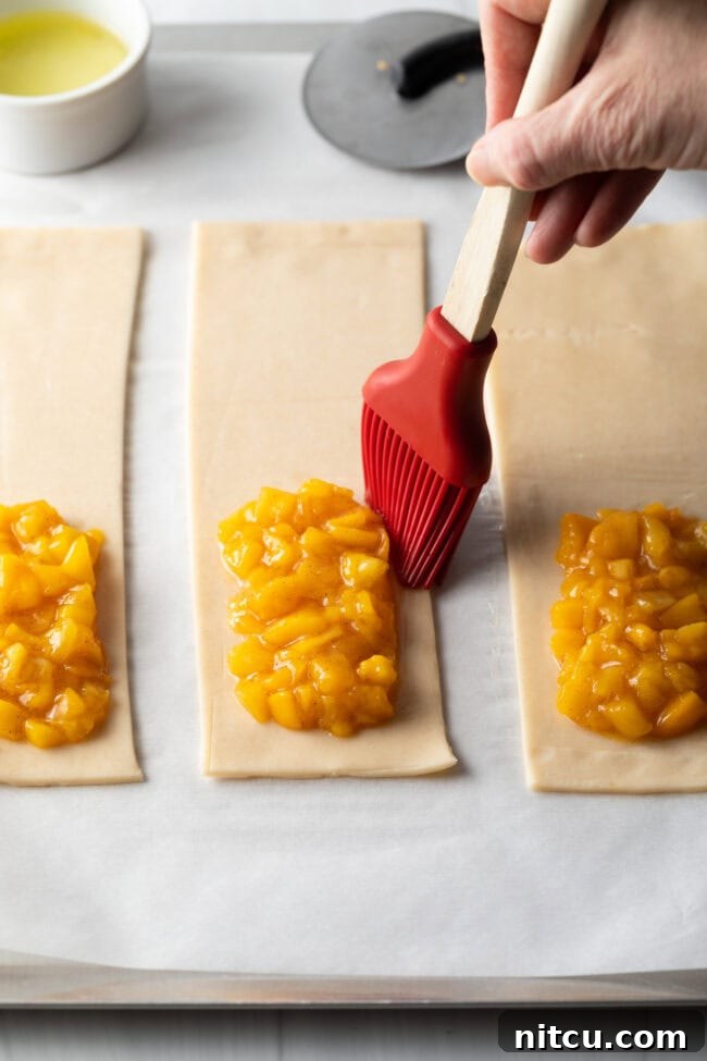 A hand using a pastry brush to apply egg white around the outside of a dollop of peach filling on a pie crust, preparing to seal the hand pie.