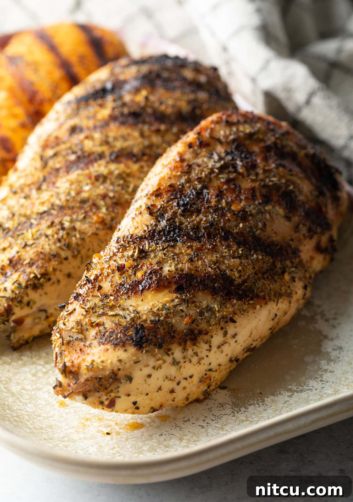 Top down view two whole chicken breasts seasoned and grilled, resting on a white plate.