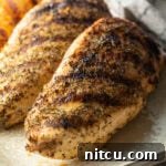 Top down view two whole chicken breasts seasoned and grilled, resting on a white plate.
