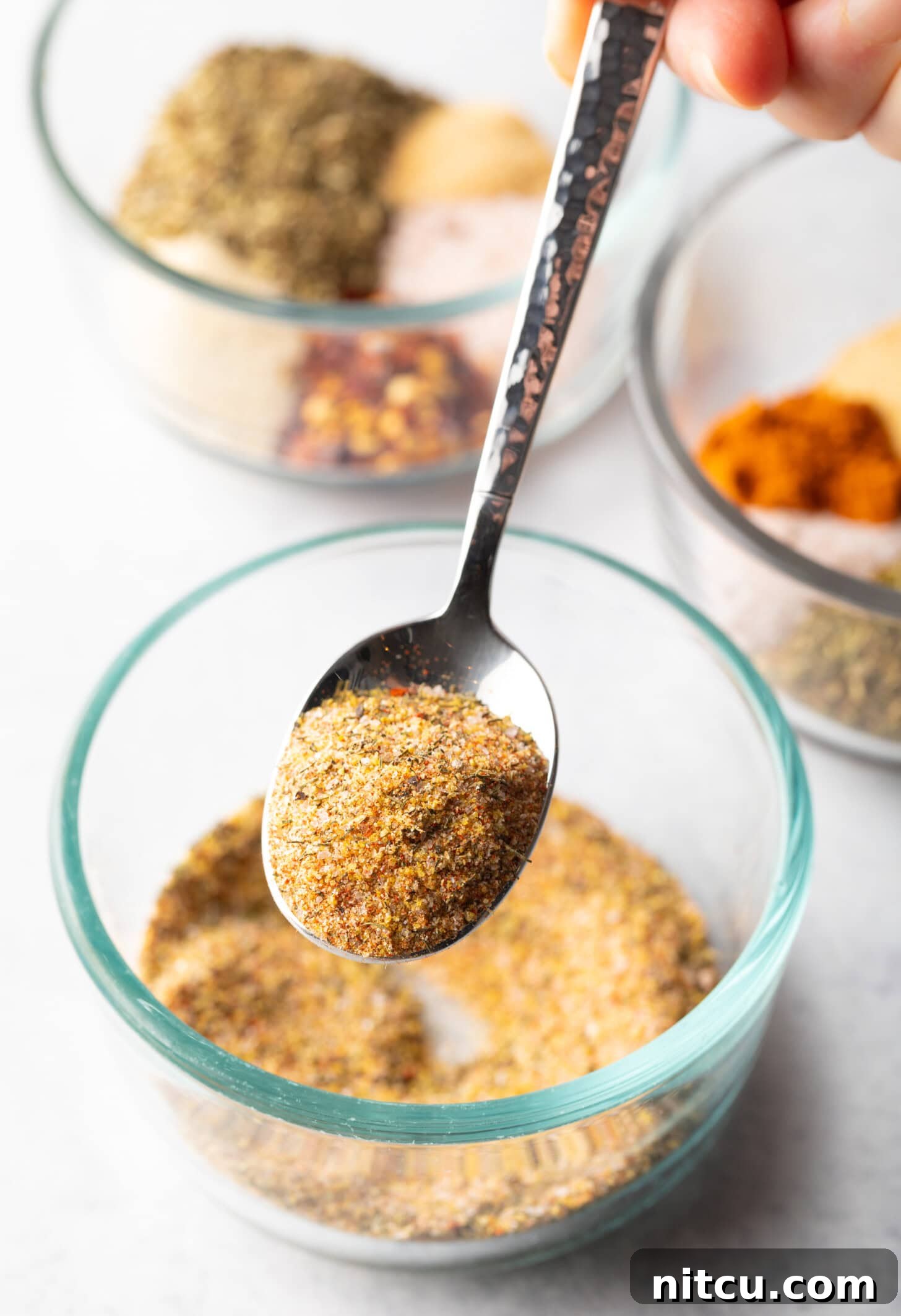 Metal spoon above glass bowl with spice blend. In the spoon is a scoop of seasoning, held to camera.