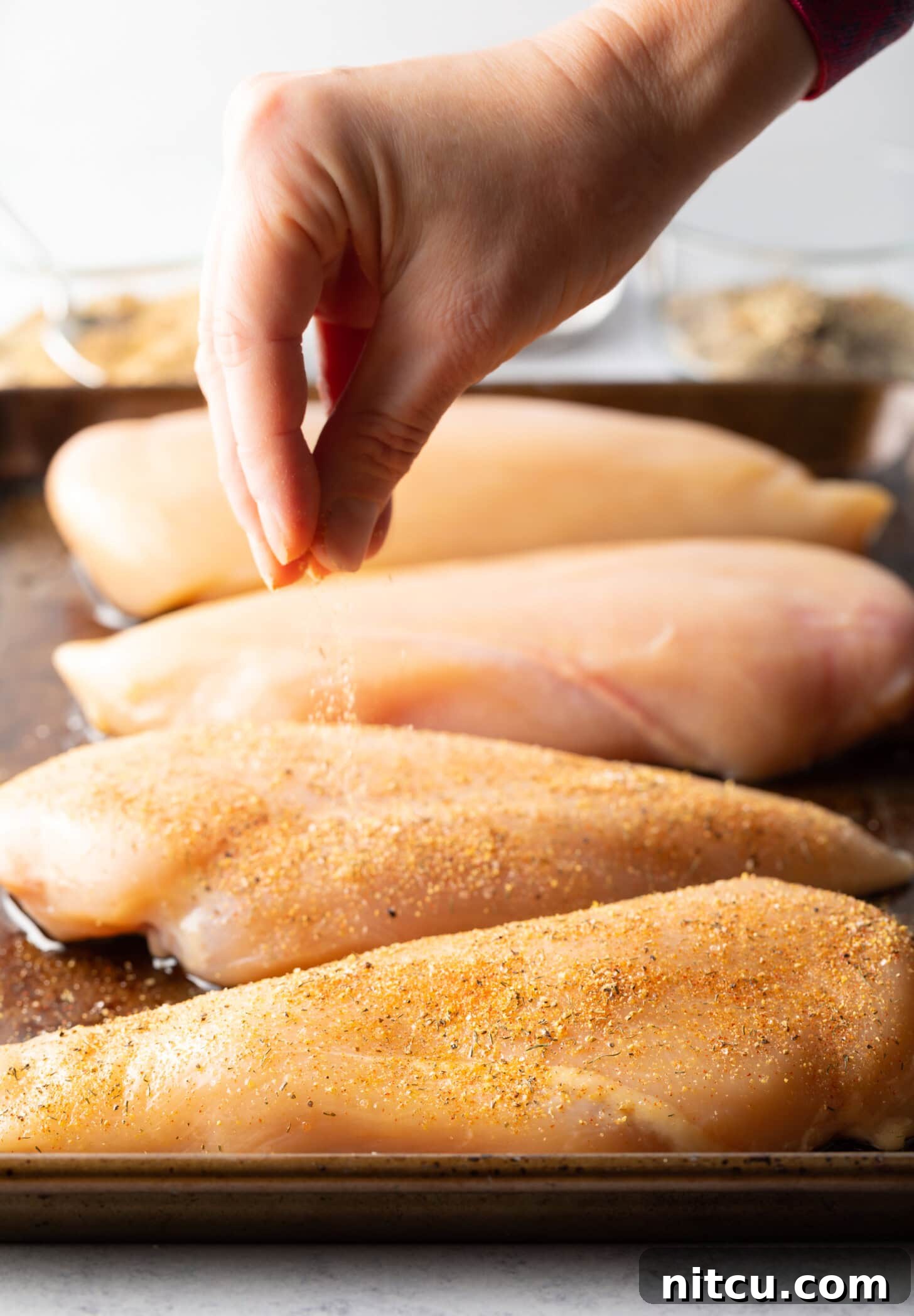 Hand sprinkling seasonings over raw chicken pieces laying out on a metal baking sheet.