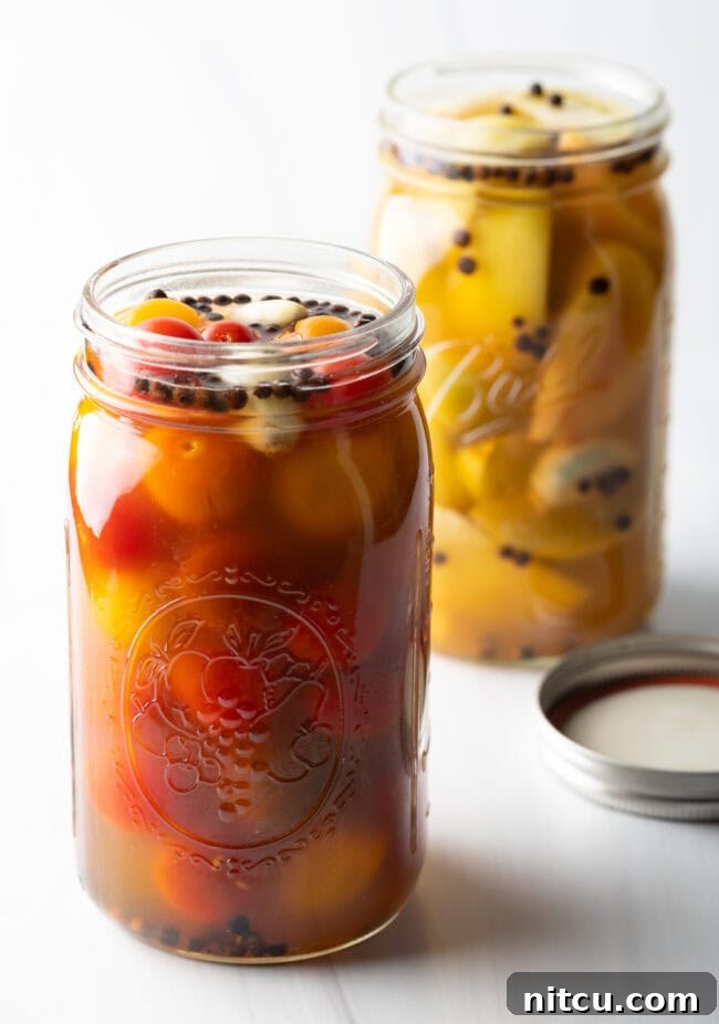 Marinated pickled cherry tomatoes in a large glass jar in the foreground. Marinated pickled green tomatoes in a large glass jar in the backrground.