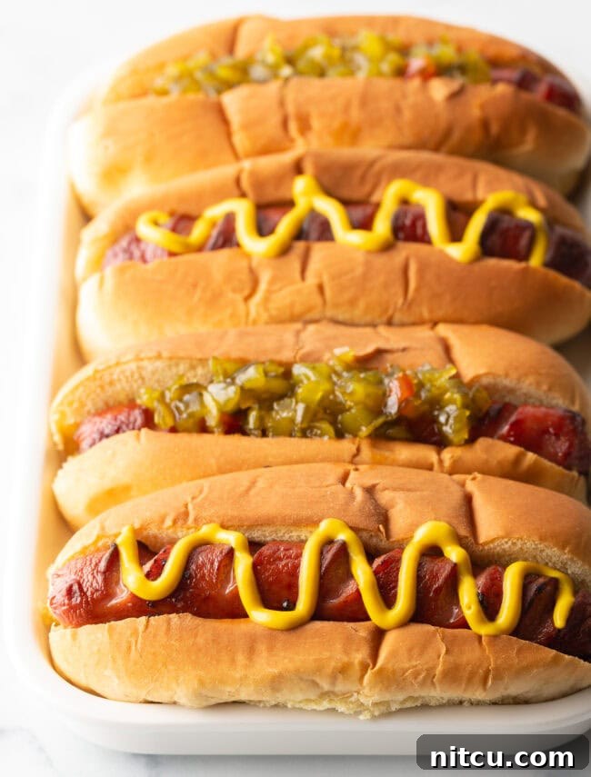 Four hotdogs in buns placed side by side on a white rectangle plate. The toppings alternative between relish and zigzag of mustard.