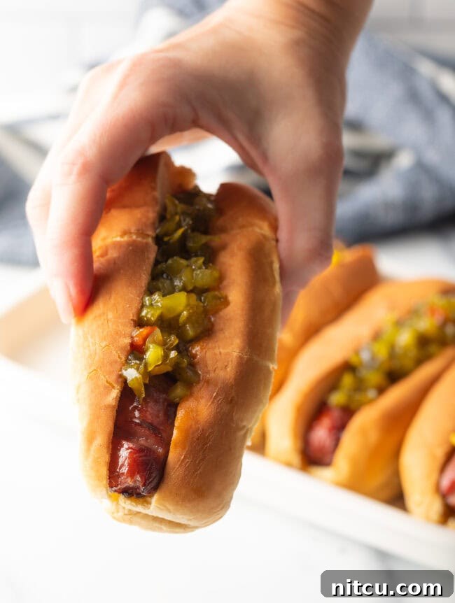 Hand holding air fried hot dog in bun dressed with relish, towards camera.