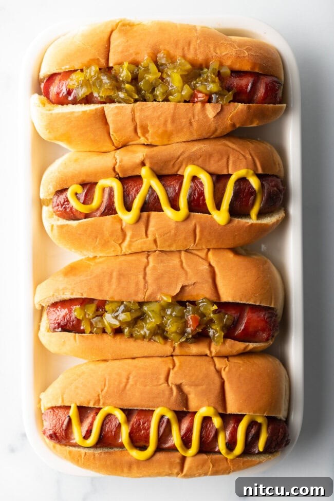 Top down view. Four hotdogs in buns placed side by side on a white rectangle plate. The toppings alternative between relish and zigzag of mustard.