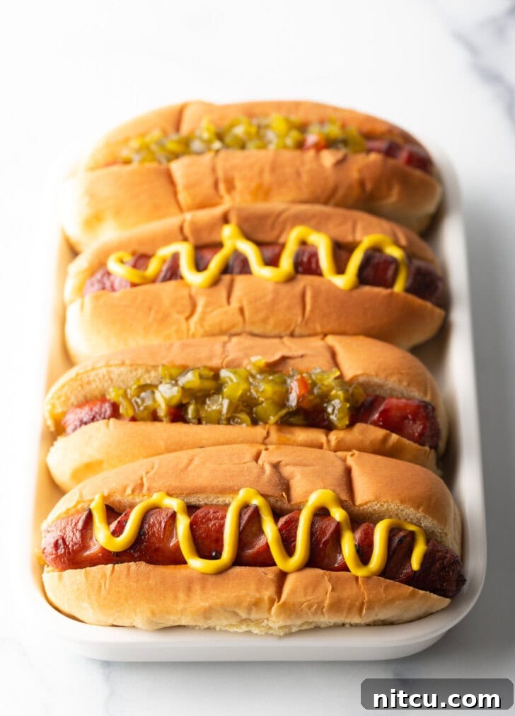 Four hotdogs in buns placed side by side on a white rectangle plate. The toppings alternative between relish and zigzag of mustard.